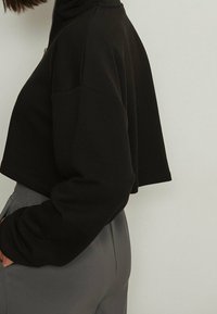 Schwarzes, kurzes Sweatshirt mit hohem Kragen und durchgehendem Reißverschluss, das lockere Ärmel und eine weiche Textur aufweist. Kombiniert mit grauen, elastischen Hosen.