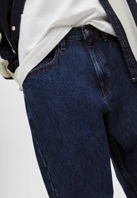 Jeans de mezclilla azul oscuro con una cintura ajustada, detalles de costura visibles y una textura ligeramente desgastada. Combinados con una camiseta blanca y una camisa azul marino.