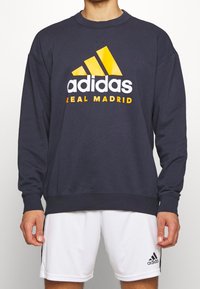 Sudadera azul marino con cuello redondo, con un gran logo en blanco y amarillo de "adidas" y el texto "REAL MADRID" impreso en el pecho.