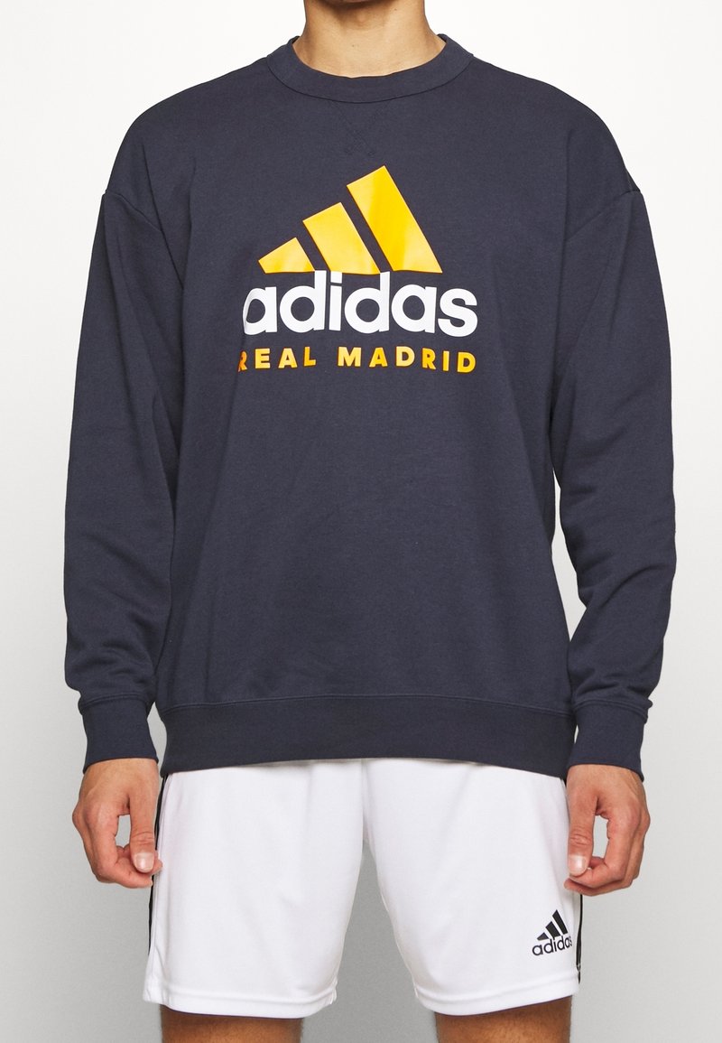 Sudadera azul marino con cuello redondo, con un gran logo en blanco y amarillo de "adidas" y el texto "REAL MADRID" impreso en el pecho.