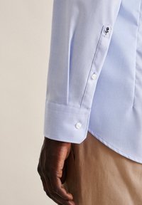 Lyseblå skjorte med lange ermer som har knappedekorert mansjett, hvite knapper og en liten svart rose brodering på ermet.
