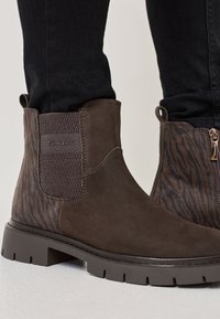 Brązowe botki za kostkę o gładkim zamszowym wykończeniu, z fakturowanymi elastycznymi panelami bocznymi oraz kontrastowym, ciemniejszym wzorem zebry na obcasie. Gruba podeszwa.