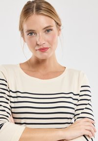 Camicia beige a strisce con linee orizzontali blu scuro, colletto rotondo e maniche a tre quarti. Tessuto a maglia morbido, vestibilità rilassata e design semplice.