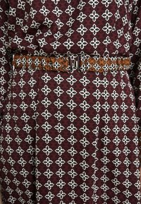Robe en tissu à motifs bordeaux, avec des motifs floraux blancs et une ceinture tressée marron avec un détail de boucle métallique.
