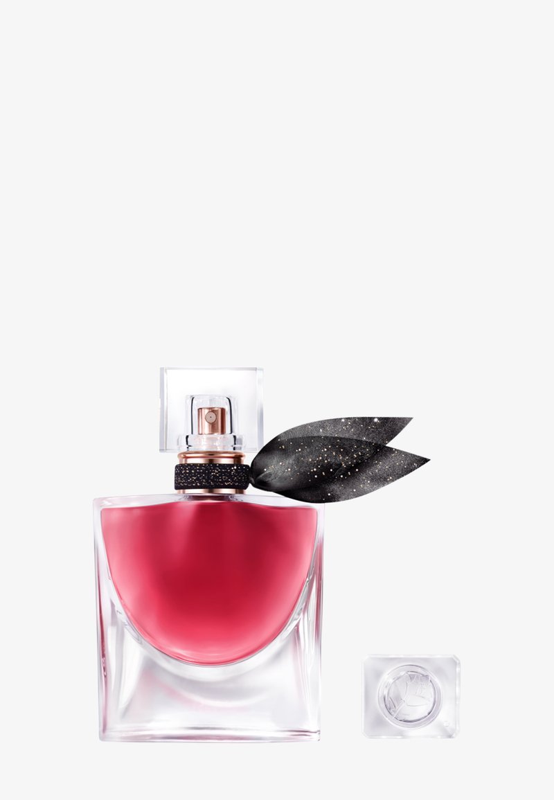 Flacon de parfum en verre transparent avec un liquide rose profond, un nœud en tissu noir avec des taches dorées au niveau du col, et un bouchon carré transparent placé à côté.