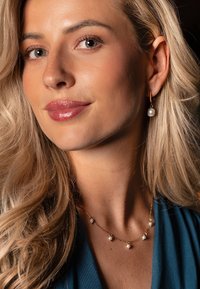 Collier chaîne en or avec petites perles et boucles d'oreilles pendantes assorties en perles. Le modèle a de longs cheveux blonds et porte un haut turquoise.