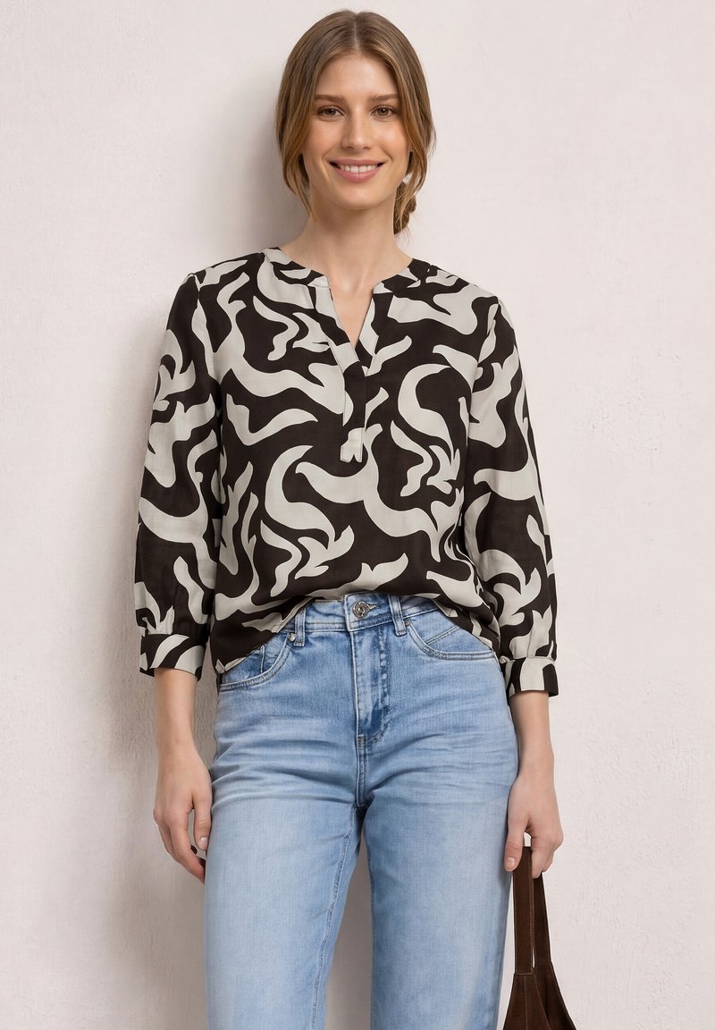 Jeune femme souriante, portant un chemisier à motif abstrait noir et blanc et un jean bleu clair, tenant un sac marron à ses côtés.