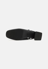 Zapato de tacón alto negro con suela de goma texturizada, detalles en la correa y tacón cuadrado. La parte inferior muestra un patrón de relieve para mayor agarre.