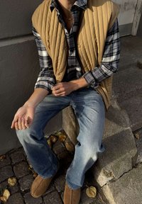 Inimene istub kivitrepil, kandes pruunikat tikandiga vest, ruudulist särk, siniseid teksapükse ja pruune kingi, sügislehed munakiviteel.