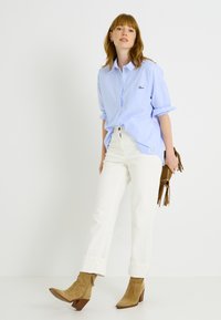 Camisa azul claro de botones con mangas remangadas, pantalones blancos de talle alto y botines de ante beige; sosteniendo un bolso marrón con flecos.