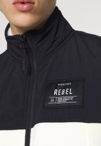 Zwart-wit ritsjack met hoge kraag. Voorzien van een rechthoekige patch met "REBEL" en andere tekst. Soepele textuur.