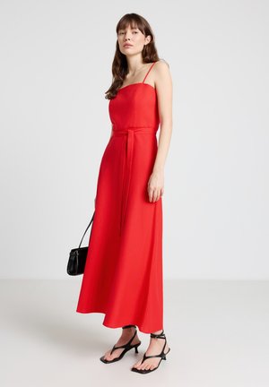 Donna che indossa un vestito lungo rosso senza maniche con spalline sottili, sandali neri con cinturini e tacco, e tiene una piccola borsa nera.