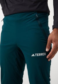 Pantalons de sport turquoise à la coupe ajustée, dotés d'une poche zippée sur le côté et du logo blanc "TERREX" sur la cuisse. Texture de tissu lisse.