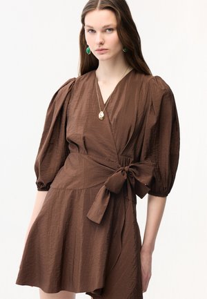 Ung kvinde iført en brun omslagskjole med puffede ærmer og en stor bundet sløjfe i taljen, accessoriseret med grønne øreringe og en guldfarvet vedhæng.