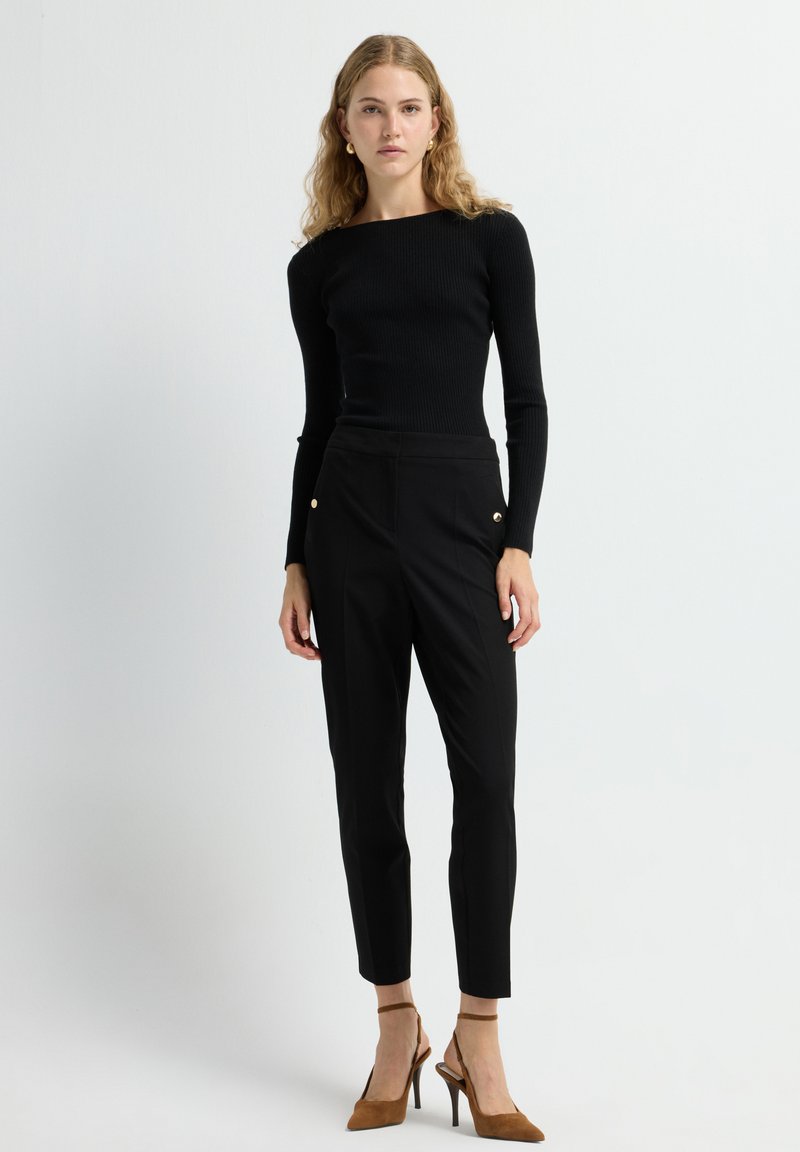 Zwarte geribbelde lange mouw top gecombineerd met zwarte tailored broek met gouden knoopaccenten, en tan suède hoge hakken.