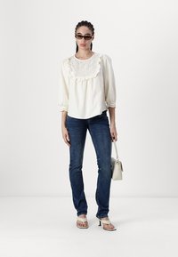 Blouse blanche avec des détails en œillets et un col volanté, assortie à un jean bleu. Les accessoires comprennent des lunettes de soleil et un sac à main léger.