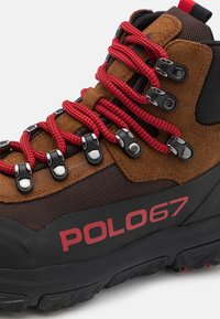 Bota de senderismo de ante marrón y malla con cordones rojos, ojales de metal y marca POLO67. Presenta una suela de goma negra texturizada para un buen agarre.
