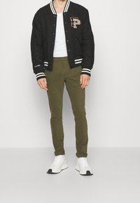Svart collegejacka med vita ränder, beige bokstavspatch, olivgröna slim-fit byxor och vita sneakers med svarta accenter.
