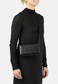Donna che indossa un vestito nero senza maniche sopra una maglietta nera a maniche lunghe, e porta una borsa a tracolla in pelle nera con logo dorato.