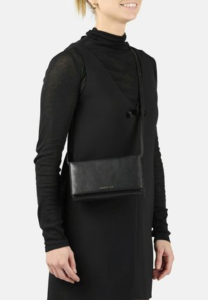 Donna che indossa un vestito nero senza maniche sopra una maglietta nera a maniche lunghe, e porta una borsa a tracolla in pelle nera con logo dorato.