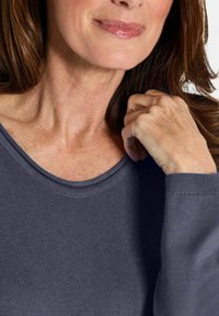 Reife Frau in einem blauen Strickpullover, die mit ihrer rechten Hand ihren Schlüsselbein berührt, mit leicht lächelnden Lippen.