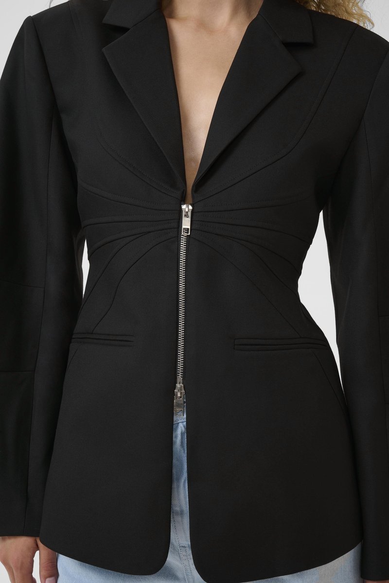 Blazer noir sur mesure avec fermeture éclair argentée à l'avant, taille cintrée, épaules structurées, porté avec un jean bleu clair par une personne.