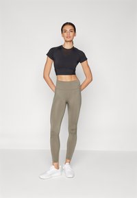 Haut de sport noir court à manches courtes, associé à un legging taille haute vert olive et des baskets blanches. Texture lisse et ajustée.