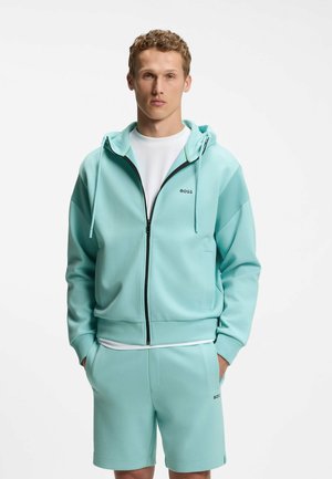 Junger Mann trägt eine hellblaue Kapuzenjacke mit Reißverschluss und passende Shorts mit einem weißen T-Shirt darunter, steht mit den Händen in den Taschen.