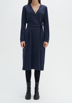 Femme portant une robe portefeuille bleu marine à manches longues, des collants noirs et des bottines noires, debout devant un fond blanc uni.