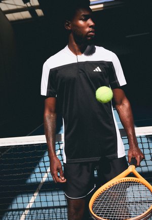 T-shirt athlétique noir et blanc à manches courtes, associé à un short noir. Tient un racket de tennis orange et une balle de tennis verte.