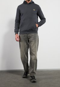 Hombre con una sudadera con capucha gris oscuro y un logo de abeja amarilla, jeans grises desgastados y zapatillas negras, de pie con una mano en el bolsillo.