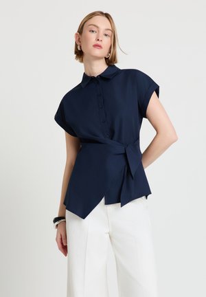 Femme aux cheveux blonds courts portant une chemise portefeuille bleu marine à boutons et un pantalon blanc à jambes larges, debout devant un fond clair uni.