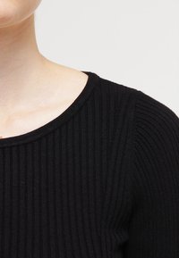 Svart ribbad stickad topp med rundad halsringning och strukturerat mönster, som visar en slät finish och en figurnära design.