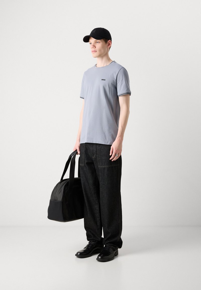 T-shirt gris clair à manches courtes avec des accents noirs, pantalon en denim foncé, casquette noire et sac de sport noir, porté avec des chaussures noires.