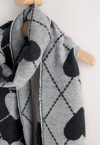 Gray scarf with a geometric pattern, featuring black hearts and dashed lines. Soft material with frayed edges, displayed on a wooden hanger.