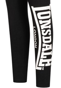 Svarta leggings i bomull med en stor vit grafisk tryck som står "Lonsdale London" längs sidan. Slät textur, åtsittande design.