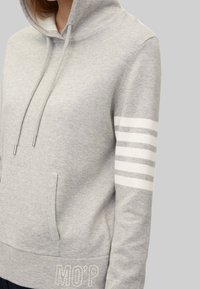 Šedá mikina s kapucí a přední kapsou, se stahovací šňůrkou a bílými pruhy na levém rukávu. Obsahuje vyšívaný detail "MO'P".