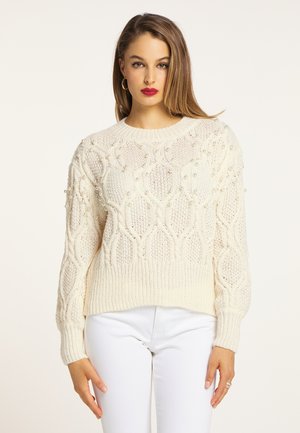 Young woman with light brown hair wearing a cream knit sweater with pearl details and white pants against a light background.