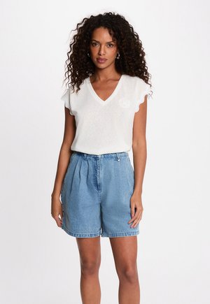 Femme aux cheveux bouclés portant un haut blanc sans manches à col en V avec des bords festonnés et un short en jean bleu clair taille haute, debout devant un fond blanc.