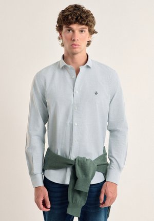 Camisa de botones a rayas en azul claro y blanco, combinada con un suéter verde atado a la cintura. Tejido suave, corte entallado.