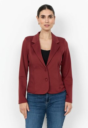 Woman with dark hair pulled back wears a maroon fitted blazer over black top and blue jeans, standing against a plain light background.