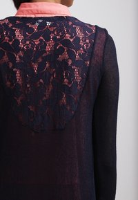 Marinblus med en genomskinlig spetsbakpanel och en korallfärgad krage. Har blommönster och ett texturerat, lätt tyg.