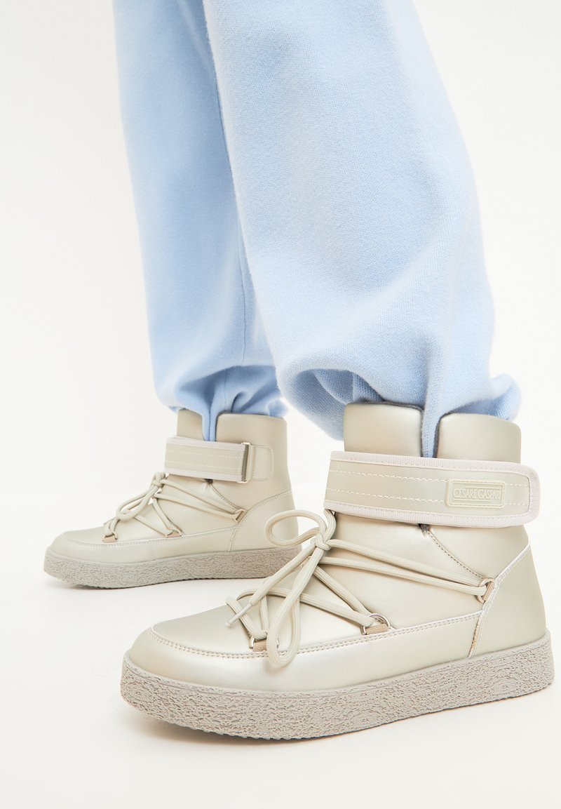 Lichtbeige enkelboots met een soepel lederen bovenwerk, verstelbare riem, decoratieve veters en een gestructureerde rubberen zool. Gecombineerd met blauwe joggers.