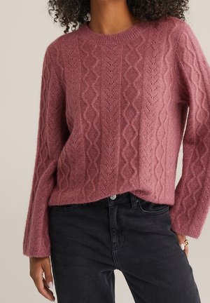 Femme portant un pull en maille torsadée texturée mauve à manches longues, associé à un jean noir taille haute et une bague en or à son doigt.