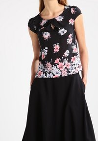 Svart blommig topp med korta puffärmar, prydd med rosa och vita blommor, rund halsringning samt en nyckelhållsdetalj framtill.