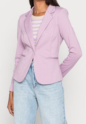 Frau trägt ein tailliertes Fliederblazer über einem beigen und lila gestreiften Oberteil, kombiniert mit hellblauen hochtaillierten Jeans vor weißem Hintergrund.