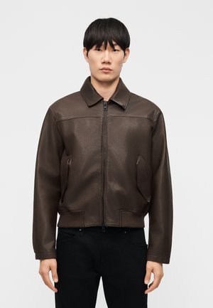 Uomo che indossa una giacca in pelle marrone scuro con cerniera e colletto, e pantaloni neri, in piedi davanti a uno sfondo chiaro e uniforme.