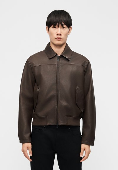 Homme portant une veste en cuir marron foncé à fermeture éclair avec col et un pantalon noir, debout devant un fond clair uni.