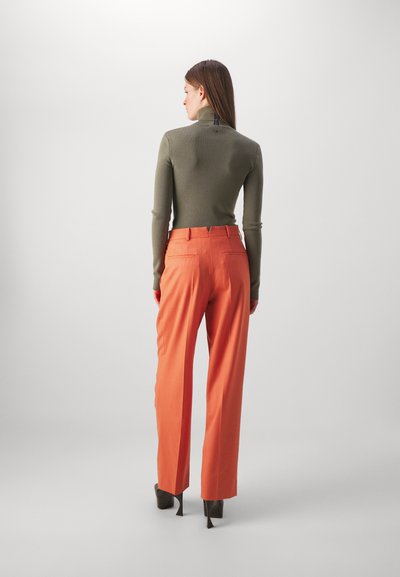 Top cu guler înalt de culoare verde măsliniu, asociat cu pantaloni largi portocalii, croiți pe măsură; textură netedă și buzunare laterale vizibile pe pantaloni. Pantofi cu toc negri.