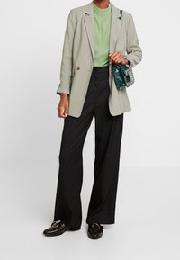 Blazer grigio chiaro, blusa verde, pantaloni neri a gamba larga e mocassini neri con dettagli in oro. Piccola borsa verde con tracolla a catena.